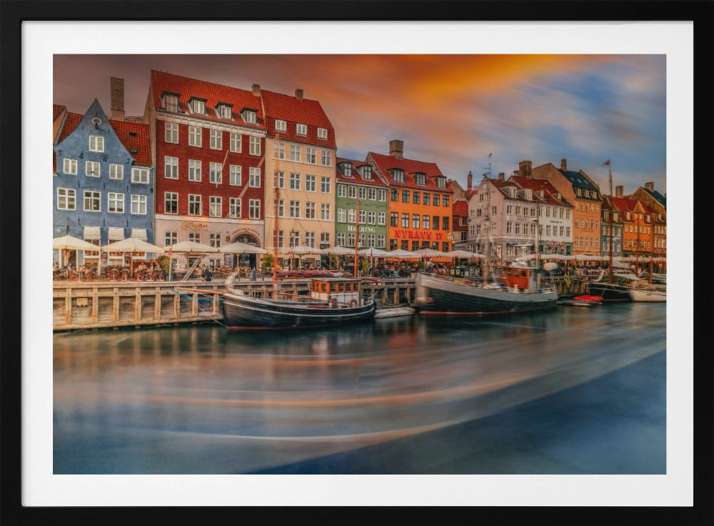 A long-exposure photograph of the colorful Nyhavn harbor in Copenhagen at sunset. Historic, brightly colored buildings line the canal, where two traditional wooden boats are moored. The sky is a dramatic blend of orange and blue, and the water is blurred, showing streaks of reflected light. Decor