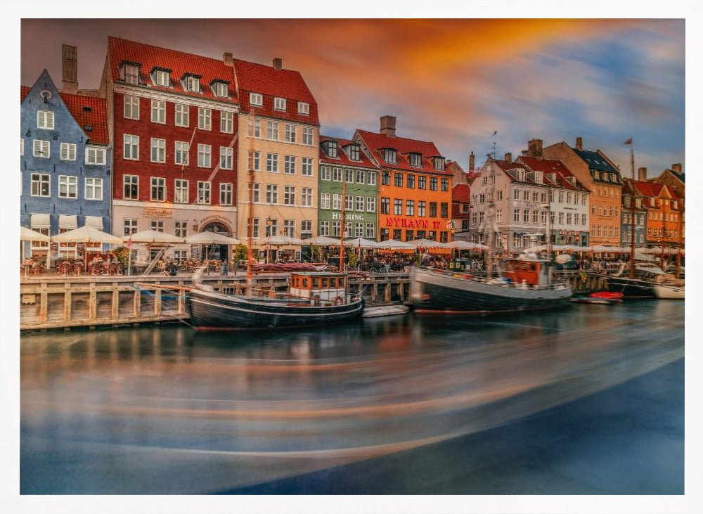 A long-exposure photograph of the colorful Nyhavn harbor in Copenhagen at sunset. Historic, brightly colored buildings line the canal, where two traditional wooden boats are moored. The sky is a dramatic blend of orange and blue, and the water is blurred, showing streaks of reflected light. Decor