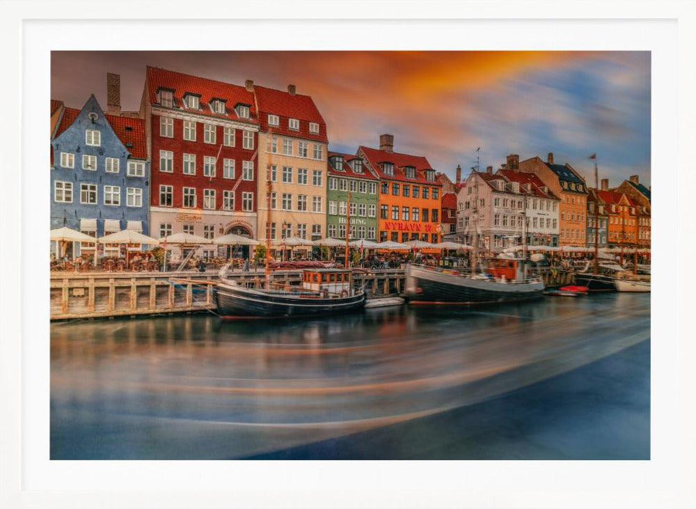 A long-exposure photograph of the colorful Nyhavn harbor in Copenhagen at sunset. Historic, brightly colored buildings line the canal, where two traditional wooden boats are moored. The sky is a dramatic blend of orange and blue, and the water is blurred, showing streaks of reflected light. Decor