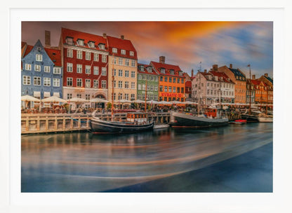 A long-exposure photograph of the colorful Nyhavn harbor in Copenhagen at sunset. Historic, brightly colored buildings line the canal, where two traditional wooden boats are moored. The sky is a dramatic blend of orange and blue, and the water is blurred, showing streaks of reflected light. Decor