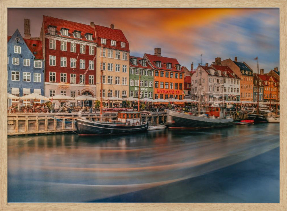 A long-exposure photograph of the colorful Nyhavn harbor in Copenhagen at sunset. Historic, brightly colored buildings line the canal, where two traditional wooden boats are moored. The sky is a dramatic blend of orange and blue, and the water is blurred, showing streaks of reflected light. Decor