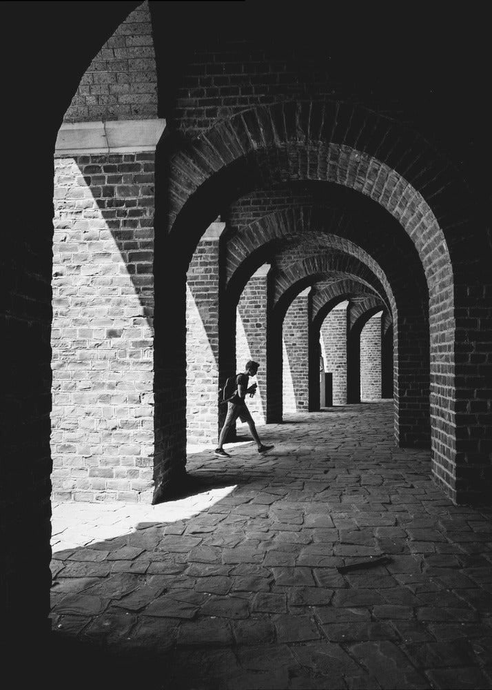 A dramatic black and white photograph of a person walking through a long brick arcade. Sunlight creates strong, contrasting shadows, highlighting the repeating arches that recede into the darkness, creating a powerful sense of depth and perspective. Decor