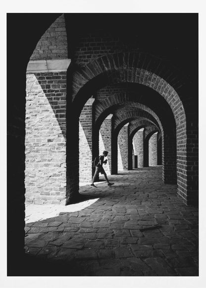 A dramatic black and white photograph of a person walking through a long brick arcade. Sunlight creates strong, contrasting shadows, highlighting the repeating arches that recede into the darkness, creating a powerful sense of depth and perspective. Decor