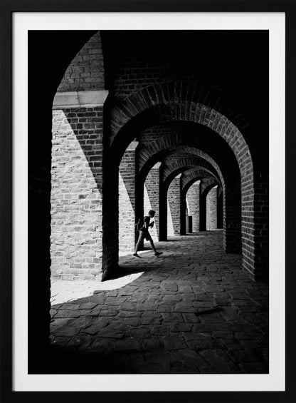 A dramatic black and white photograph of a person walking through a long brick arcade. Sunlight creates strong, contrasting shadows, highlighting the repeating arches that recede into the darkness, creating a powerful sense of depth and perspective. Decor