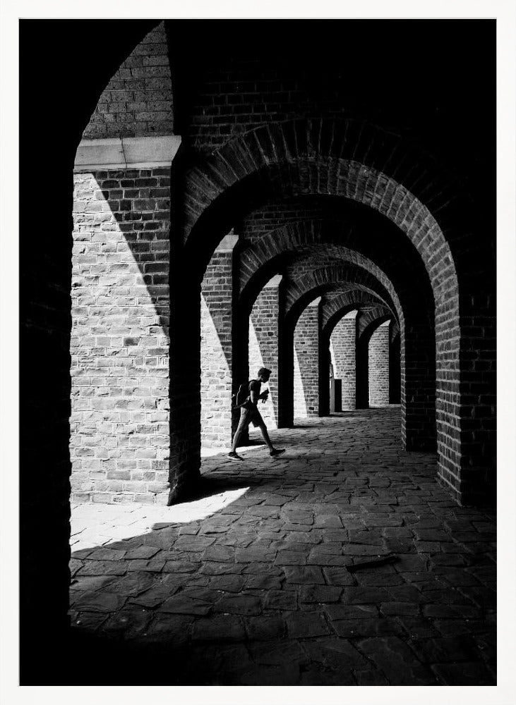 A dramatic black and white photograph of a person walking through a long brick arcade. Sunlight creates strong, contrasting shadows, highlighting the repeating arches that recede into the darkness, creating a powerful sense of depth and perspective. Decor