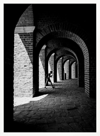 A dramatic black and white photograph of a person walking through a long brick arcade. Sunlight creates strong, contrasting shadows, highlighting the repeating arches that recede into the darkness, creating a powerful sense of depth and perspective. Decor