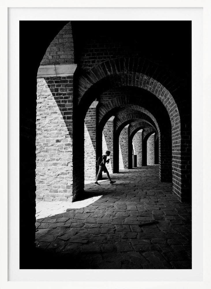 A dramatic black and white photograph of a person walking through a long brick arcade. Sunlight creates strong, contrasting shadows, highlighting the repeating arches that recede into the darkness, creating a powerful sense of depth and perspective. Decor