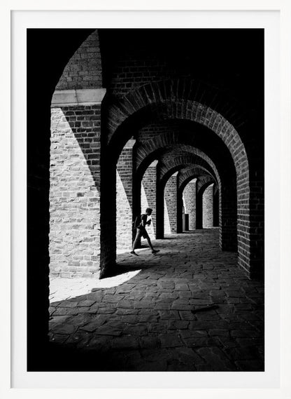 A dramatic black and white photograph of a person walking through a long brick arcade. Sunlight creates strong, contrasting shadows, highlighting the repeating arches that recede into the darkness, creating a powerful sense of depth and perspective. Decor