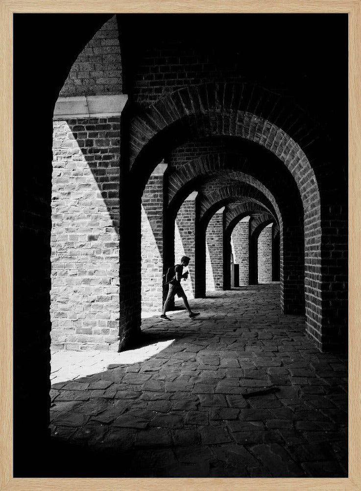 A dramatic black and white photograph of a person walking through a long brick arcade. Sunlight creates strong, contrasting shadows, highlighting the repeating arches that recede into the darkness, creating a powerful sense of depth and perspective. Decor