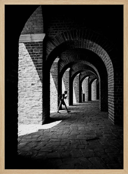 A dramatic black and white photograph of a person walking through a long brick arcade. Sunlight creates strong, contrasting shadows, highlighting the repeating arches that recede into the darkness, creating a powerful sense of depth and perspective. Decor