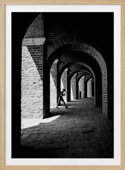 A dramatic black and white photograph of a person walking through a long brick arcade. Sunlight creates strong, contrasting shadows, highlighting the repeating arches that recede into the darkness, creating a powerful sense of depth and perspective. Decor