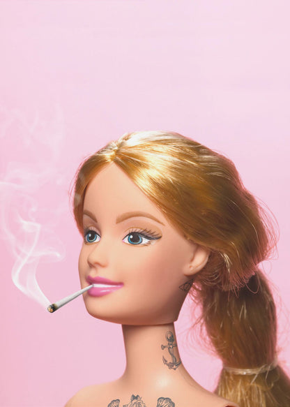 A close-up photograph of a blonde doll, resembling Barbie, with tattoos on her neck and chest, smoking a joint against a solid pink background. Smoke curls upwards from the lit cigarette in her pink-lipsticked mouth. Artwork