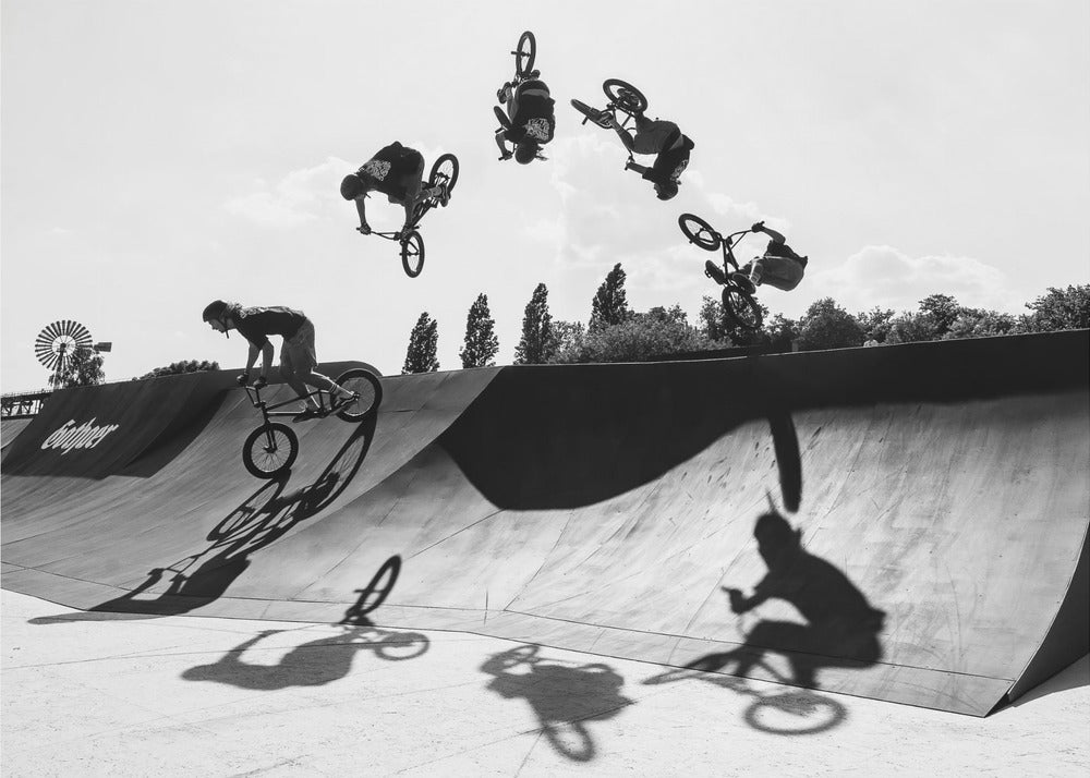 A dynamic black and white action shot showing the progression of a BMX rider doing a backflip off a large ramp. Multiple exposures capture the rider at different points in the air, creating an arc of motion against a bright sky. Strong shadows are cast on the ramp below. Print