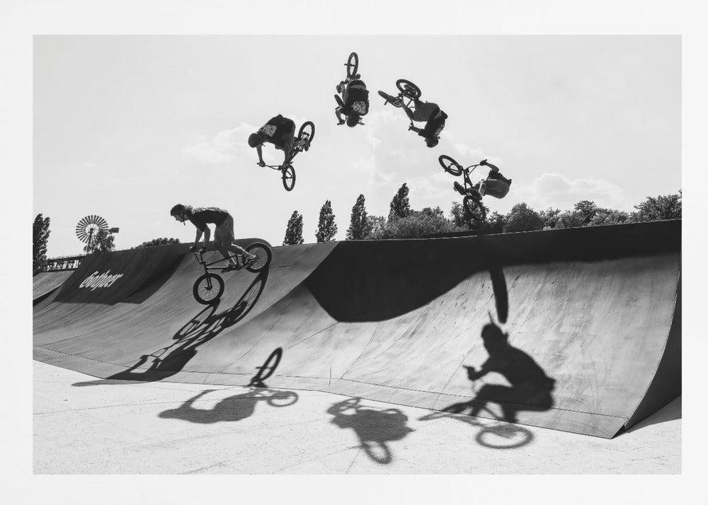 A dynamic black and white action shot showing the progression of a BMX rider doing a backflip off a large ramp. Multiple exposures capture the rider at different points in the air, creating an arc of motion against a bright sky. Strong shadows are cast on the ramp below. Print