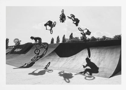 A dynamic black and white action shot showing the progression of a BMX rider doing a backflip off a large ramp. Multiple exposures capture the rider at different points in the air, creating an arc of motion against a bright sky. Strong shadows are cast on the ramp below. Print