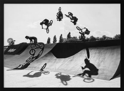A dynamic black and white action shot showing the progression of a BMX rider doing a backflip off a large ramp. Multiple exposures capture the rider at different points in the air, creating an arc of motion against a bright sky. Strong shadows are cast on the ramp below. Print