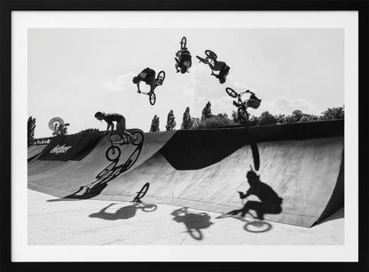 A dynamic black and white action shot showing the progression of a BMX rider doing a backflip off a large ramp. Multiple exposures capture the rider at different points in the air, creating an arc of motion against a bright sky. Strong shadows are cast on the ramp below. Print