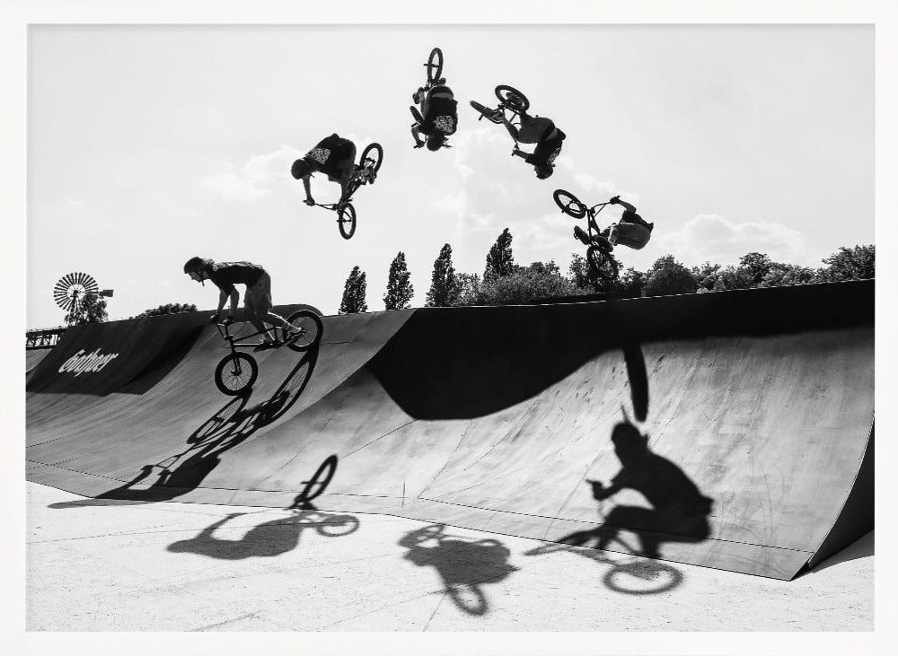 A dynamic black and white action shot showing the progression of a BMX rider doing a backflip off a large ramp. Multiple exposures capture the rider at different points in the air, creating an arc of motion against a bright sky. Strong shadows are cast on the ramp below. Print