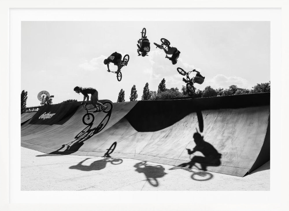 A dynamic black and white action shot showing the progression of a BMX rider doing a backflip off a large ramp. Multiple exposures capture the rider at different points in the air, creating an arc of motion against a bright sky. Strong shadows are cast on the ramp below. Print