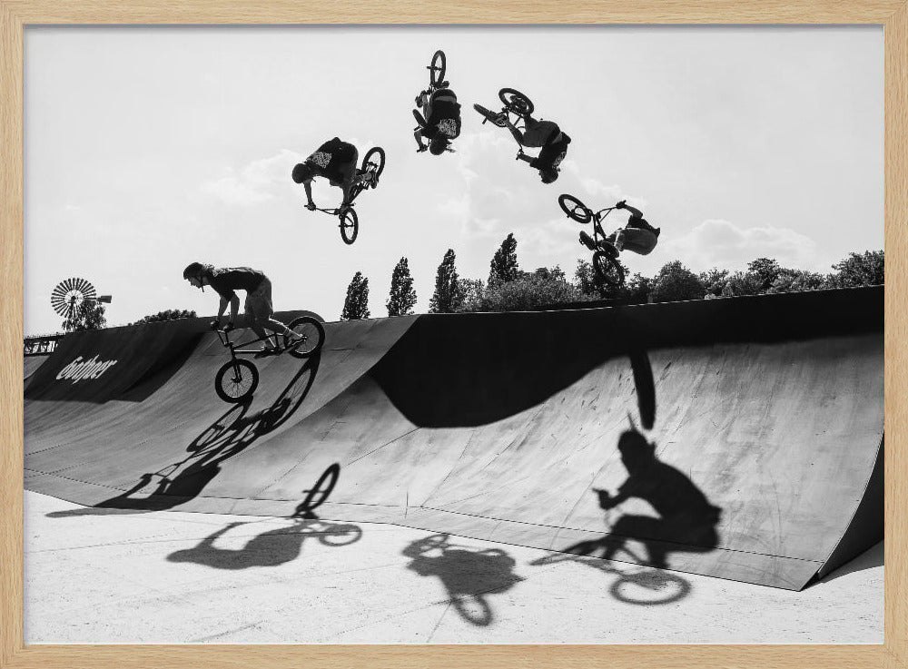 A dynamic black and white action shot showing the progression of a BMX rider doing a backflip off a large ramp. Multiple exposures capture the rider at different points in the air, creating an arc of motion against a bright sky. Strong shadows are cast on the ramp below. Print