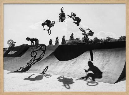 A dynamic black and white action shot showing the progression of a BMX rider doing a backflip off a large ramp. Multiple exposures capture the rider at different points in the air, creating an arc of motion against a bright sky. Strong shadows are cast on the ramp below. Print