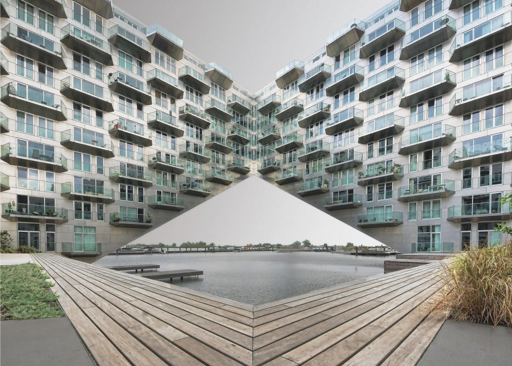 A symmetrical, mirrored photograph of a modern V-shaped apartment complex with numerous glass balconies, overlooking a wooden deck and a calm body of water under a grey sky. The image is presented within a silver picture frame. Artwork