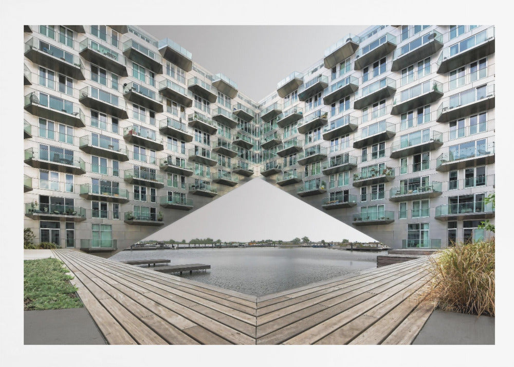 A symmetrical, mirrored photograph of a modern V-shaped apartment complex with numerous glass balconies, overlooking a wooden deck and a calm body of water under a grey sky. The image is presented within a silver picture frame. Artwork