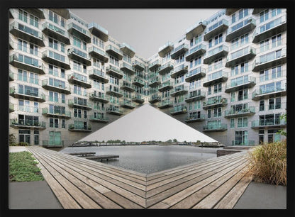 A symmetrical, mirrored photograph of a modern V-shaped apartment complex with numerous glass balconies, overlooking a wooden deck and a calm body of water under a grey sky. The image is presented within a silver picture frame. Artwork