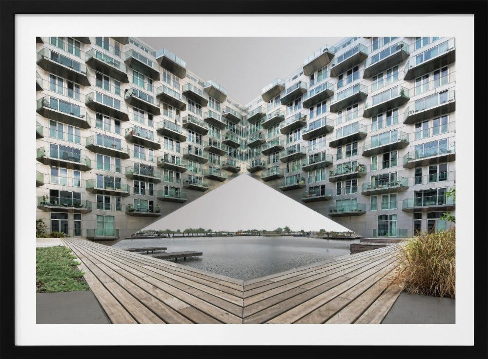 A symmetrical, mirrored photograph of a modern V-shaped apartment complex with numerous glass balconies, overlooking a wooden deck and a calm body of water under a grey sky. The image is presented within a silver picture frame. Artwork
