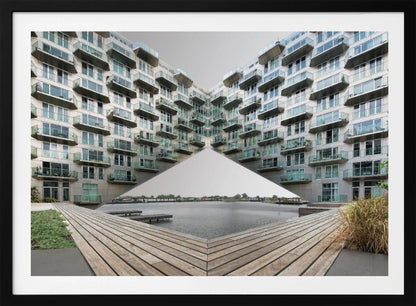 A symmetrical, mirrored photograph of a modern V-shaped apartment complex with numerous glass balconies, overlooking a wooden deck and a calm body of water under a grey sky. The image is presented within a silver picture frame. Artwork