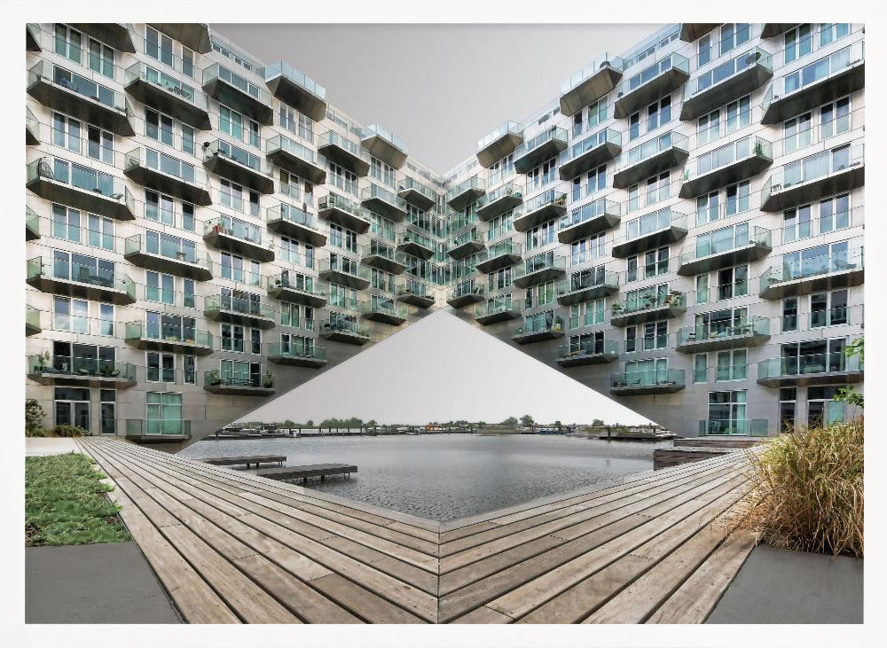 A symmetrical, mirrored photograph of a modern V-shaped apartment complex with numerous glass balconies, overlooking a wooden deck and a calm body of water under a grey sky. The image is presented within a silver picture frame. Artwork