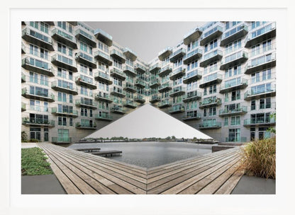 A symmetrical, mirrored photograph of a modern V-shaped apartment complex with numerous glass balconies, overlooking a wooden deck and a calm body of water under a grey sky. The image is presented within a silver picture frame. Artwork