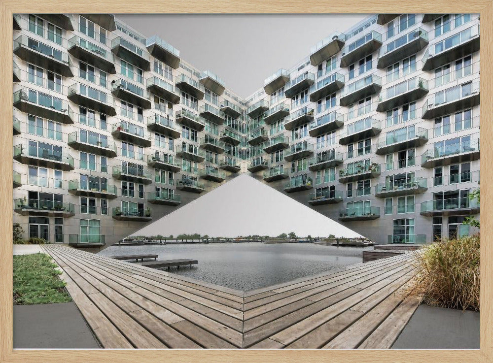 A symmetrical, mirrored photograph of a modern V-shaped apartment complex with numerous glass balconies, overlooking a wooden deck and a calm body of water under a grey sky. The image is presented within a silver picture frame. Artwork
