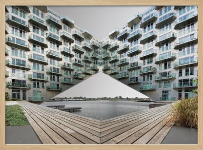 A symmetrical, mirrored photograph of a modern V-shaped apartment complex with numerous glass balconies, overlooking a wooden deck and a calm body of water under a grey sky. The image is presented within a silver picture frame. Artwork