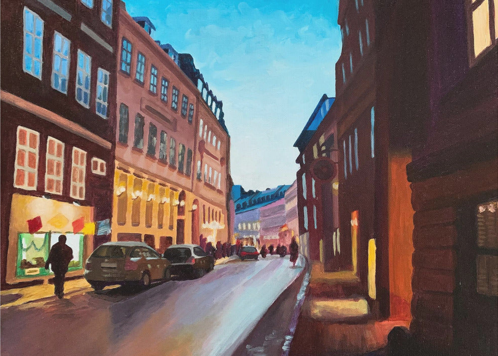 An impressionistic painting of a European city street at dusk, shown in a silver frame. The sky is a bright, clear blue, contrasting with the deep brown and maroon buildings. Warm yellow light glows from shop windows and street lamps, reflecting on the wet-looking street where cars are parked. Decor