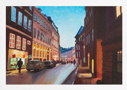 An impressionistic painting of a European city street at dusk, shown in a silver frame. The sky is a bright, clear blue, contrasting with the deep brown and maroon buildings. Warm yellow light glows from shop windows and street lamps, reflecting on the wet-looking street where cars are parked. Decor