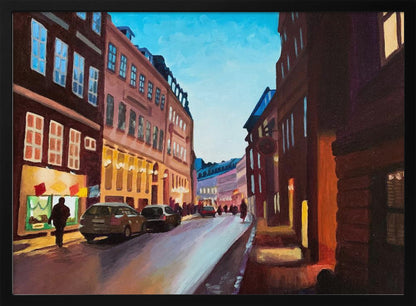 An impressionistic painting of a European city street at dusk, shown in a silver frame. The sky is a bright, clear blue, contrasting with the deep brown and maroon buildings. Warm yellow light glows from shop windows and street lamps, reflecting on the wet-looking street where cars are parked. Decor