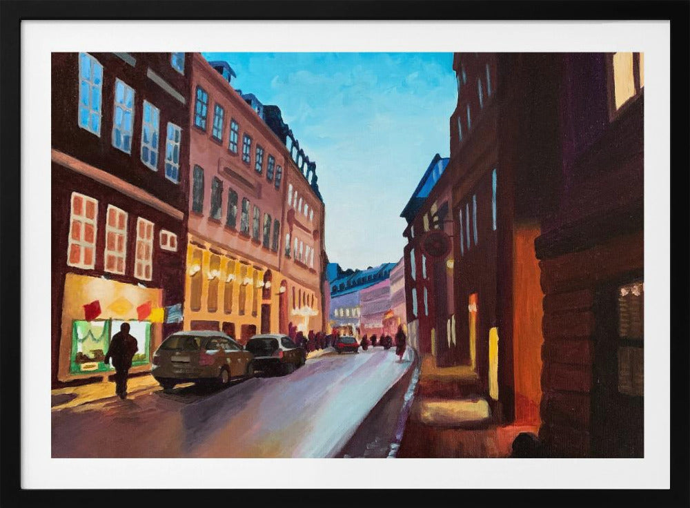 An impressionistic painting of a European city street at dusk, shown in a silver frame. The sky is a bright, clear blue, contrasting with the deep brown and maroon buildings. Warm yellow light glows from shop windows and street lamps, reflecting on the wet-looking street where cars are parked. Decor