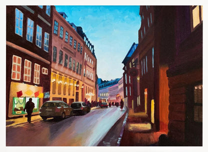 An impressionistic painting of a European city street at dusk, shown in a silver frame. The sky is a bright, clear blue, contrasting with the deep brown and maroon buildings. Warm yellow light glows from shop windows and street lamps, reflecting on the wet-looking street where cars are parked. Decor