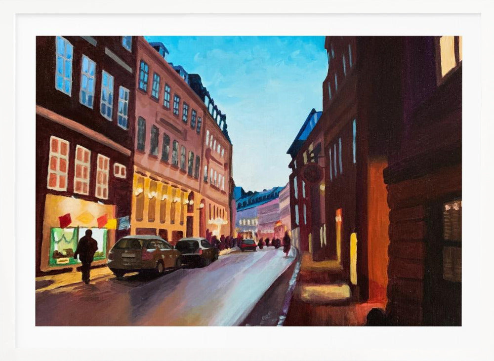 An impressionistic painting of a European city street at dusk, shown in a silver frame. The sky is a bright, clear blue, contrasting with the deep brown and maroon buildings. Warm yellow light glows from shop windows and street lamps, reflecting on the wet-looking street where cars are parked. Decor