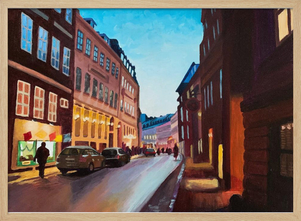 An impressionistic painting of a European city street at dusk, shown in a silver frame. The sky is a bright, clear blue, contrasting with the deep brown and maroon buildings. Warm yellow light glows from shop windows and street lamps, reflecting on the wet-looking street where cars are parked. Decor