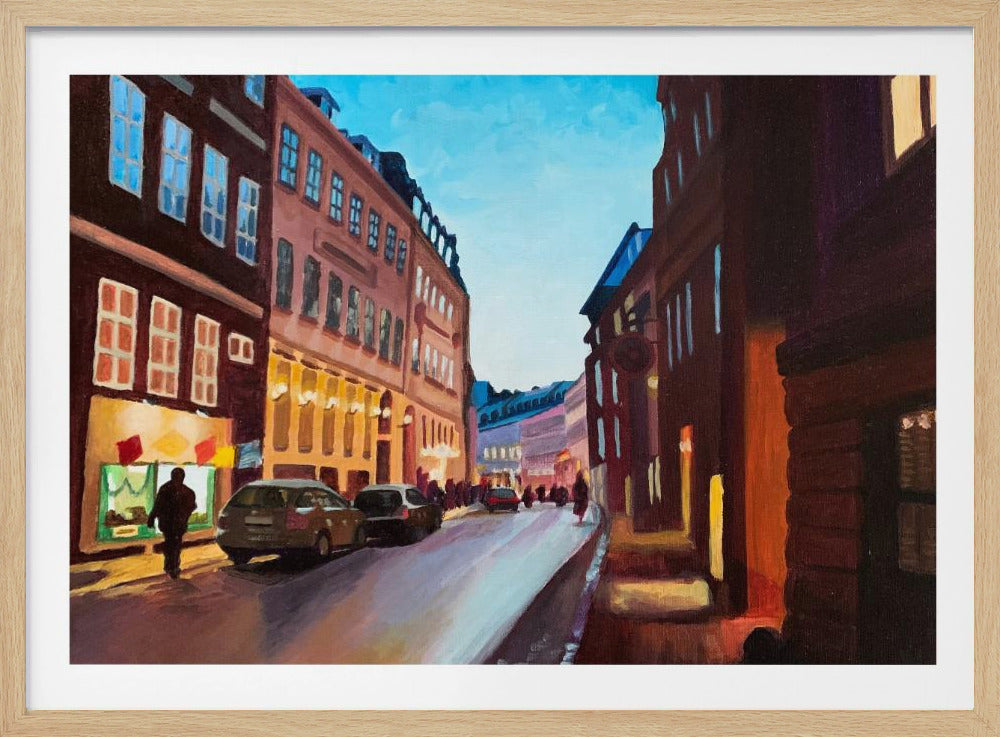 An impressionistic painting of a European city street at dusk, shown in a silver frame. The sky is a bright, clear blue, contrasting with the deep brown and maroon buildings. Warm yellow light glows from shop windows and street lamps, reflecting on the wet-looking street where cars are parked. Decor