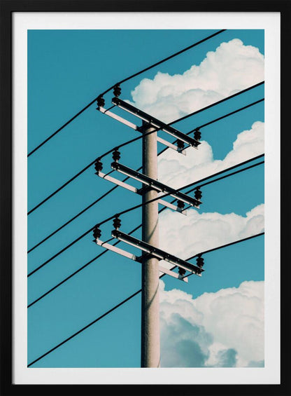 A low-angle photograph of a concrete utility pole with black power lines stretching diagonally across the frame against a vibrant blue sky with fluffy white clouds. Decor