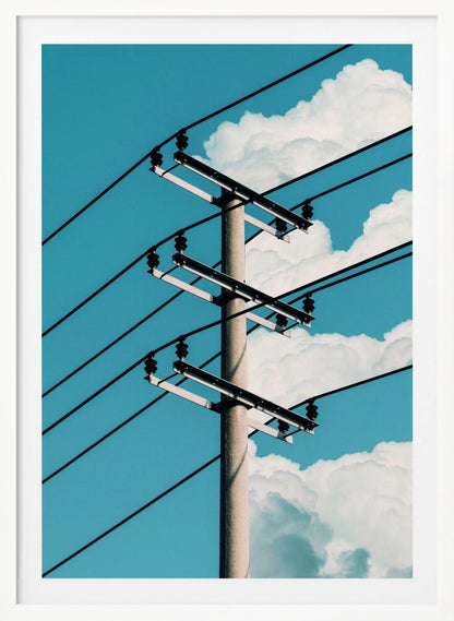A low-angle photograph of a concrete utility pole with black power lines stretching diagonally across the frame against a vibrant blue sky with fluffy white clouds. Decor