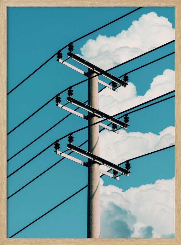 A low-angle photograph of a concrete utility pole with black power lines stretching diagonally across the frame against a vibrant blue sky with fluffy white clouds. Decor