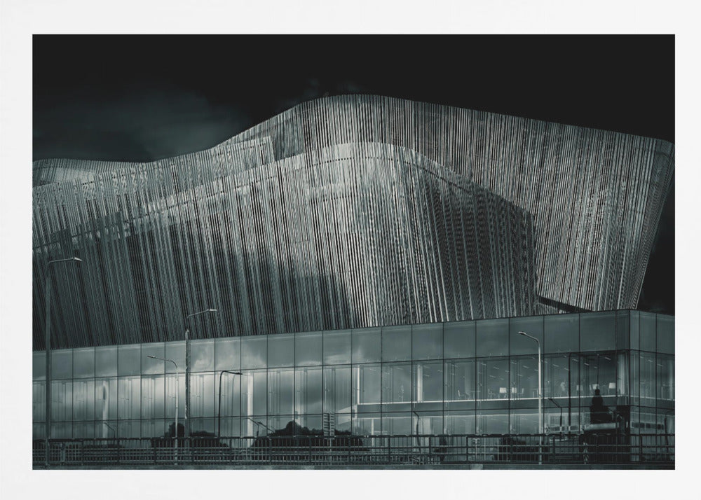 A dramatic black and white photograph of a modern building with a unique, wave-like facade made of vertical fins. The lower level is made of glass, reflecting the sky and streetlights. The entire image is presented in a silver frame. Artwork