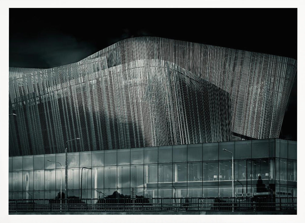 A dramatic black and white photograph of a modern building with a unique, wave-like facade made of vertical fins. The lower level is made of glass, reflecting the sky and streetlights. The entire image is presented in a silver frame. Artwork