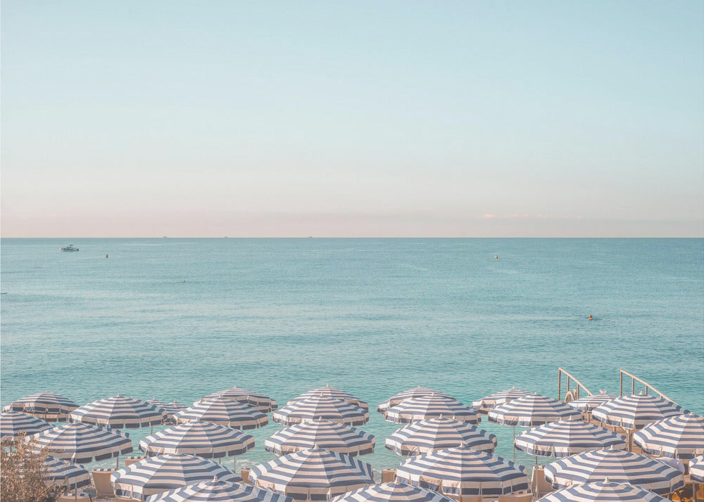 A high-angle photograph of a beach packed with blue and white striped umbrellas, with the turquoise ocean stretching out to a pale blue sky in the background. The entire image is enclosed in a silver frame. Poster