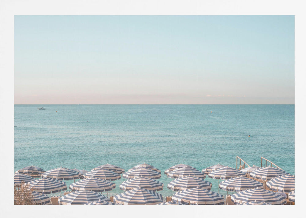 A high-angle photograph of a beach packed with blue and white striped umbrellas, with the turquoise ocean stretching out to a pale blue sky in the background. The entire image is enclosed in a silver frame. Poster