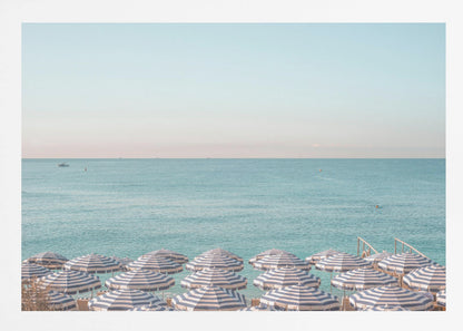 A high-angle photograph of a beach packed with blue and white striped umbrellas, with the turquoise ocean stretching out to a pale blue sky in the background. The entire image is enclosed in a silver frame. Poster